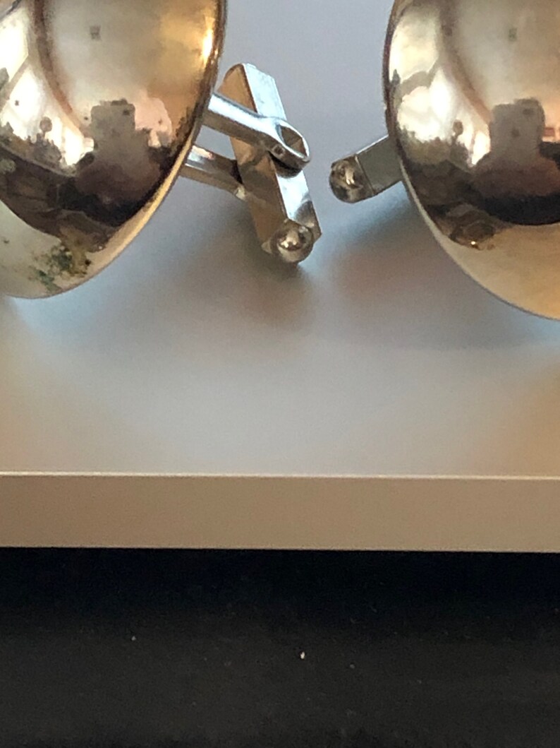 May include: A pair of silver cufflinks with a round, gold-toned design. The cufflinks are connected by a silver bar.