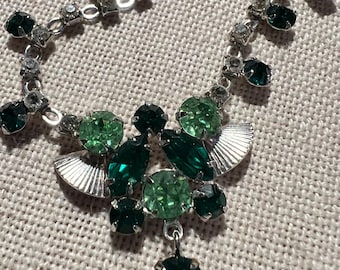 May include: A silver necklace with a green rhinestone pendant. The pendant is made up of several green rhinestones in different shades of green. The necklace is made of silver chain with small clear rhinestones.