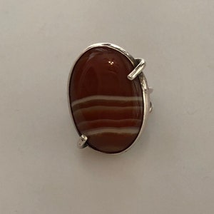 Pu&ograve; includere: Un anello d'argento con una grande pietra ovale di corniola. La pietra ha strisce bianche.