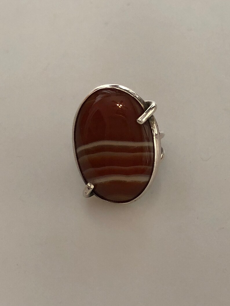 Pu&ograve; includere: Un anello d'argento con una grande pietra ovale di corniola. La pietra ha strisce bianche.