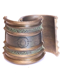 Op de afbeelding: Een gouden metalen armband met een decoratief ontwerp. De armband heeft een getextureerd oppervlak en een band met een cirkelvormig ontwerp in het midden. De armband is aan één kant open en heeft aan de andere kant een scharnier.