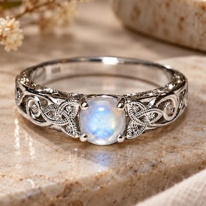 May include: A silver-toned ring featuring a central round, iridescent moonstone cabochon. The band is crafted with intricate, openwork metal detailing that displays repeating knotwork patterns on both sides of the gemstone setting.