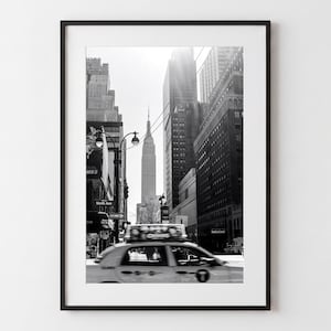 May include: A black and white framed photograph of a New York City street scene featuring a yellow taxi cab in motion, a tall skyscraper resembling the Empire State Building in the distance, street lamps, and surrounding urban architecture.