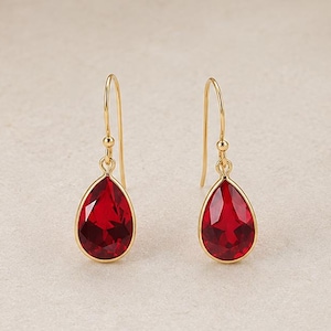 May include: A pair of gold drop earrings featuring faceted, teardrop-shaped red stones. Each earring is suspended from a thin gold hook wire, with the vibrant red gemstone encased in a polished gold setting that highlights the clear, reflective facets.