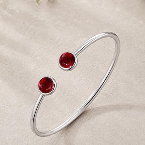 May include: A silver-toned open cuff bracelet featuring two polished, circular red stones at each end. The metal band is smooth and slender, creating a minimalist circular shape that remains open at the front for easy placement on the wrist.