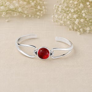 May include: A silver-toned cuff bracelet featuring a central, circular, deep red gemstone. The band is constructed with a split design that curves inward toward the stone, creating an open, polished metal frame that encircles the wrist.