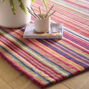 May include: A rectangular area rug featuring a vibrant pattern of multi-coloured vertical stripes in shades of red, blue, yellow, green, and white. A white ceramic cup filled with pencils sits atop a book titled happy starts at home.