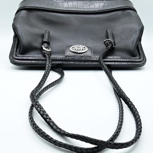 Puede incluir: Un bolso tote de cuero negro con una correa de cuero trenzada. El bolso tiene una hebilla plateada y un logotipo plateado en la parte delantera.