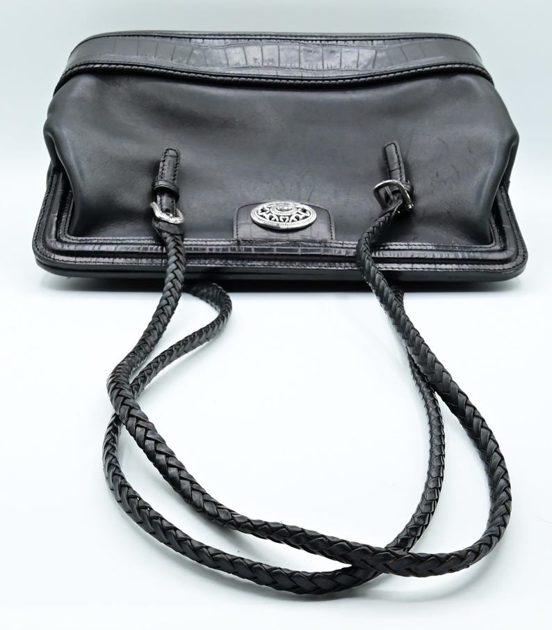 Puede incluir: Un bolso tote de cuero negro con una correa de cuero trenzada. El bolso tiene una hebilla plateada y un logotipo plateado en la parte delantera.