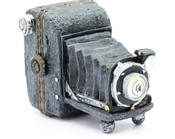 May include: A vintage-style black camera with a silver lens and a decorative detail on the side. The camera has a textured surface and a worn look.