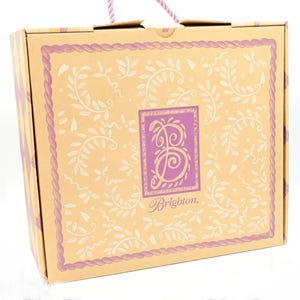 Puede incluir: Una caja de cart&oacute;n marr&oacute;n con un dise&ntilde;o floral morado y blanco. La caja tiene un asa y el texto "Brighton." impreso en la parte delantera.