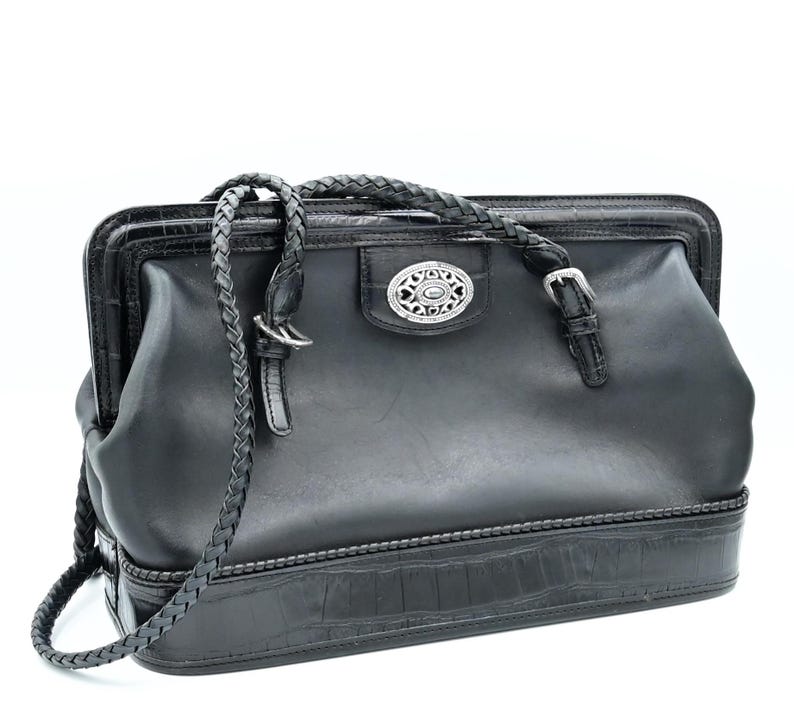 Puede incluir: Bolso de mano de cuero negro con una correa trenzada y una hebilla plateada. El bolso tiene una base con estampado de cocodrilo y un detalle decorativo plateado en la parte delantera.