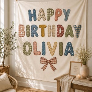 May include: A cream-colored fabric wall hanging featuring the text HAPPY BIRTHDAY OLIVIA in large, multi-colored, patterned letters. Each letter displays a unique design, including gingham, floral, and star prints. A plaid bow is centered below.