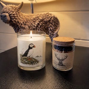 Peut inclure: Une bougie en verre transparent avec une mèche allumée et une étiquette illustrée d'un macareux, à côté d'un petit bocal en verre rempli d'allumettes à bout noir, avec un couvercle en liège et une étiquette montrant un cerf et le texte HT DESIGN.