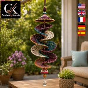 Op de afbeelding: Een hangend spiraalornament gemaakt van veelkleurig haakgaren in tinten bordeaux, groenblauw en beige. De structuur is voorzien van een centrale koord met houten kralen, een gehaakte kap aan de bovenkant en een helder gefacetteerde kristal.
