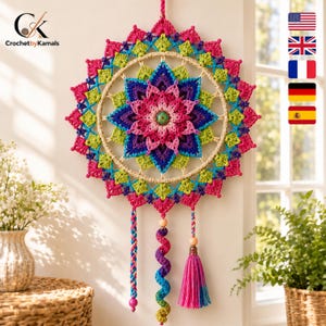 Op de afbeelding: Een ronde gehaakte wandhanger met een meerlaags mandala-ontwerp in levendige tinten roze, blauw en limoengroen. Het midden heeft een kleine decoratieve knoop en het stuk bevat drie hangende kwastjes met kralen en een spiraal.