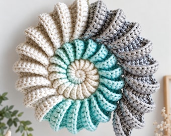 Puede incluir: Un tapiz de ganchillo circular con un diseño de concha en espiral. Los colores del hilo pasan de crema a turquesa y terminan en un plateado metálico. Una pequeña concha está unida al lazo superior para colgar el objeto.