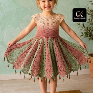 May include: A crochet dress featuring a gradient color pattern transitioning from beige at the round neckline to dusty rose and sage green at the hem. The skirt has a flared, pleated design with scalloped edges and small wooden beads.