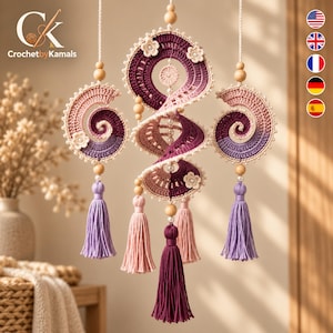 Op de afbeelding: Een hangende gehaakte mobiel met drie spiraalvormige ornamenten in tinten paars, roze en crème. De stukken zijn versierd met kleine witte kralen, houten bollen en gehaakte bloemen, afgewerkt met lange, bungelende kwastjes van garen.