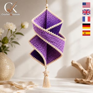 Puede incluir: Un adorno colgante en espiral hecho de tela de ganchillo morada con bordes festoneados de color crema. La estructura cuelga de una cadena dorada con una cuenta blanca y presenta un dije de estrella y una borla de color crema en la base.