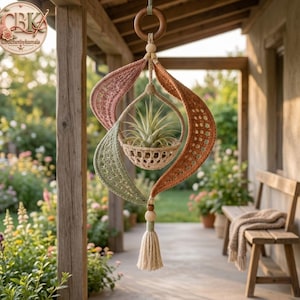 May include: A hanging crochet plant holder featuring a central basket containing a spiky air plant. The structure is composed of three curved, textured panels in muted pink, sage green, and terracotta orange, accented with wooden beads and a tassel.