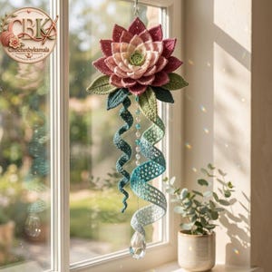 May include: A hanging crochet decoration featuring a large, multi-layered lotus flower in shades of pink, cream, and green. Below the flower, two long, spiraling crochet tendrils in teal and light blue descend alongside a string of clear beads.