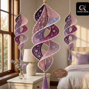 Puede incluir: Tres móviles en espiral colgantes hechos de ganchillo en tonos morado, lavanda y rosa. Cada pieza presenta intrincados patrones de encaje, un borde festoneado con pequeños pompones blancos, una cuenta de madera y una borla morada a juego.
