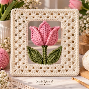 May include: A square crochet granny square frame in off-white yarn featuring a three-dimensional pink tulip with green leaves in the center. The text Crochetbykamals is printed in brown at the bottom center of the square crochet piece.