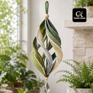 Puede incluir: Un adorno de ganchillo en espiral colgante que presenta tonos alternos de verde oscuro, verde claro, gris y crema. La estructura está rematada con una cuenta de madera y un lazo para colgar, y termina en la parte inferior con un cristal transparente.