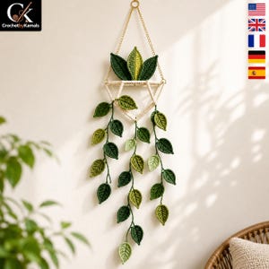 May include: A wall-hanging crochet plant featuring a geometric cream-coloured frame suspended by a gold chain. The piece includes three upright leaves and three long, trailing vines with multiple dark and light green crocheted leaves attached.