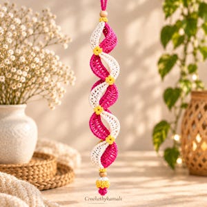 May include: A hanging crochet ornament featuring a double-helix spiral design in alternating white and magenta yarn. The structure is accented with small yellow crocheted flowers at each twist, finished with wooden beads and a yellow tassel at the base.