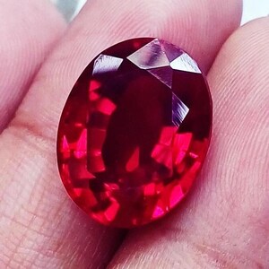 May include: A vibrant, deep red gemstone with an oval shape and a faceted cut. The stone displays a high level of clarity and brilliance, with multiple geometric facets reflecting light across its surface, creating a rich, saturated crimson color.