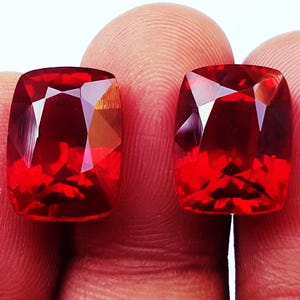 May include: Two cushion-cut gemstones with a vibrant, deep red colour and faceted surfaces that reflect light. These transparent stones exhibit a clear, polished finish with sharp edges and a symmetrical shape, showcasing a brilliant red hue throughout.