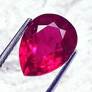 May include: A pear-shaped, faceted ruby gemstone with a deep, vibrant red colour. The gemstone is held by a pair of metal tweezers, showcasing its clear, polished surface and precise geometric cuts that reflect light across its translucent body.