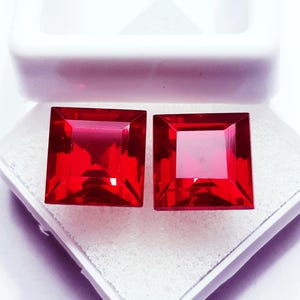 Puede incluir: Dos piedras preciosas de color rojo vibrante con corte cuadrado y superficies facetadas, dispuestas una al lado de la otra. Las piedras están colocadas dentro de un joyero blanco abierto con un inserto de espuma blanca texturizada.