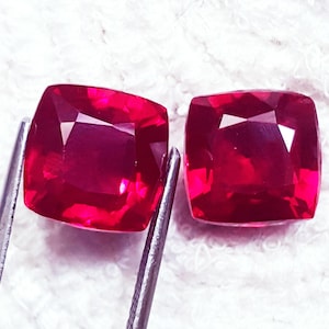 May include: Two cushion-cut, vibrant red gemstones with faceted surfaces. One gemstone is held by silver-toned metal tweezers, while the other sits adjacent to it. The stones exhibit a deep, saturated crimson hue and a high-gloss, reflective finish.
