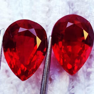 May include: Two pear-shaped, faceted red gemstones with a brilliant cut. The stones exhibit a deep, vibrant crimson hue and are held by silver-toned metal tweezers, showcasing their reflective surfaces and precise geometric facets.