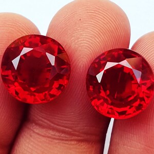 May include: Two round, faceted, vibrant red gemstones with a brilliant cut. Each stone displays a deep crimson hue and multiple triangular facets that reflect light, creating a sparkling appearance across the surface of the circular gems.