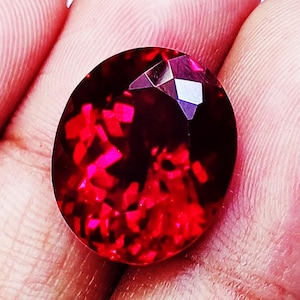 Pode incluir: Uma pedra preciosa de cor vermelho profundo e formato oval com uma superfície facetada que reflete a luz em padrões carmesim brilhantes. A pedra apresenta um acabamento polido e uma qualidade translúcida, exibindo uma cor rica e saturada.
