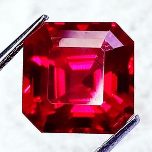 May include: A vibrant, deep red gemstone with an emerald-cut shape, featuring sharp, clean facets that reflect light. The stone is held by metallic silver-toned tweezers at the top left and bottom right corners, showcasing its clarity and color.