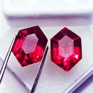 Può includere: Due vivaci gemme rosse sfaccettate esagonalmente con bordi netti e un aspetto trasparente ad alta chiarezza. Le pietre mostrano profonde tonalità cremisi e sono tenute da pinzette metalliche color argento che ne esaltano il taglio.