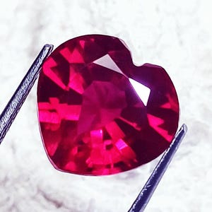 May include: A vibrant, deep red gemstone cut into a heart shape with multiple facets that reflect light. The stone is held by two thin, dark metal prongs on the left and right sides, showcasing its translucent quality and intense crimson color.