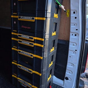 May include: A vertical stack of black and yellow DeWalt tool storage cases secured with a green bungee cord. The cases feature labels reading PLUMBING, SNAP-ON, HARDWARE, ELECTRICAL, and HAND TOOLS, with additional branding for Wera visible.
