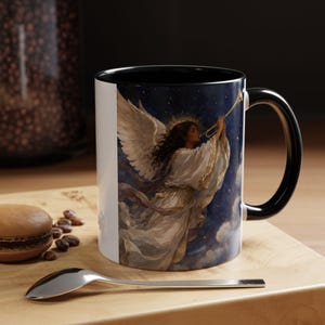 Puede incluir: Una taza de cerámica blanca con interior y asa negros, que presenta una ilustración impresa de un ángel con grandes alas blancas tocando una trompeta dorada sobre un cielo azul oscuro estrellado lleno de nubes y un halo brillante alrededor.