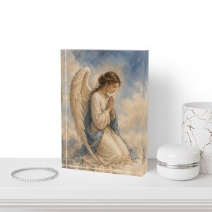 Puede incluir: Un bloque acrílico rectangular con una ilustración impresa de un ángel con grandes alas blancas, arrodillado con las manos juntas en oración. El ángel viste una túnica blanca con un manto azul sobre un fondo de nubes azules y crema.