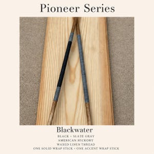 May include: Pioneer Series Blackwater sticks made of American hickory wood. One stick features a solid black waxed linen thread wrap, while the other has a slate gray accent wrap. The sticks are displayed against a light wood grain background.