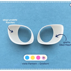 Könnte beinhalten: Zwei weiße, oval geformte Kunststoff-Augenkomponenten mit glatten Oberflächen und abgerundeten Kanten. Darunter befindet sich eine Leiste mit blauen, gelben und pinken Kreisen, einem Pluszeichen sowie dem Text "viele Farben + Größen!" und "Bastelio".