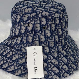 Könnte beinhalten: Ein marineblauer und beigefarbener Fischerhut mit einem sich wiederholenden Monogrammmuster. Der Hut verfügt über zwei Metallösen an der Seite und ein weißes, rechteckiges Etikett, das vorne hängt und den Text Christian Dior in Schwarz zeigt.