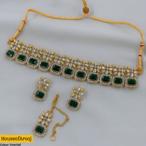 Può includere: Un set di gioielli color oro composto da una collana girocollo, due orecchini e un maang tikka. Ogni pezzo presenta pietre rettangolari verde smeraldo incorniciate da cristalli bianchi disposti in motivi floreali con un cordino dorato.