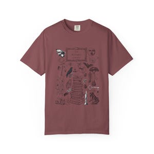 May include: A dusty rose short-sleeve t-shirt featuring a black ink-style graphic print. The design includes a stack of books, a candle, a crow, a frog, mushrooms, a moon, and the text "The Midnight Library" centered in a rectangular frame.